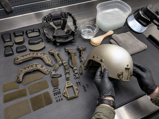 Technician inspecting a disassembled tactical helmet with pads, retention straps, rails, mounting hardware, and cleaning supplies.
